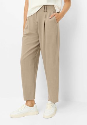 Pantalon beige plissé avec passants pour ceinture, porté avec des baskets blanches, une main dans la poche, montré de la taille aux pieds sur un fond uni.
