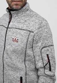 Grå zip-up jakke med et tekstureret strikkemønster, der har sorte accenter, en høj krave, og et broderet logo på brystet. Lynlåslomme på ærmet.