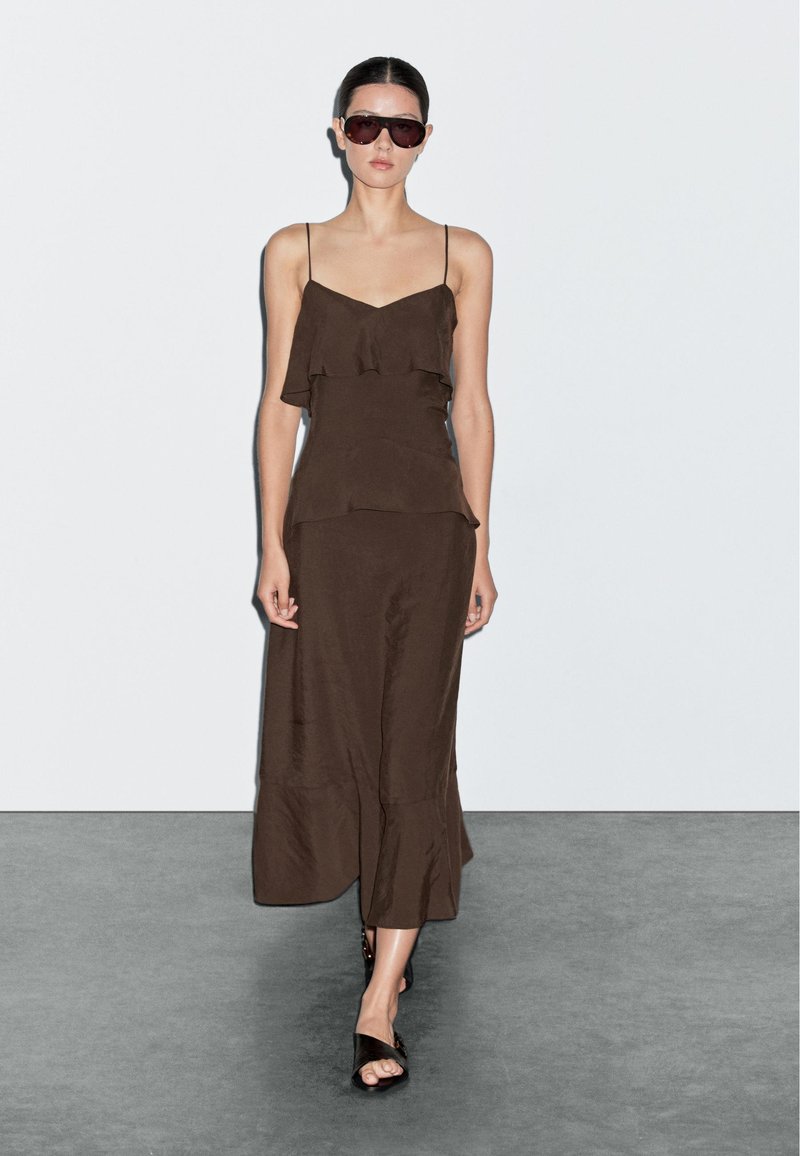 Hnědé bezramínkové šaty s volánkovými vrstvami, které sahají do půli lýtek. Na sobě má tmavé, oversize sluneční brýle a otevřené černé sandály.