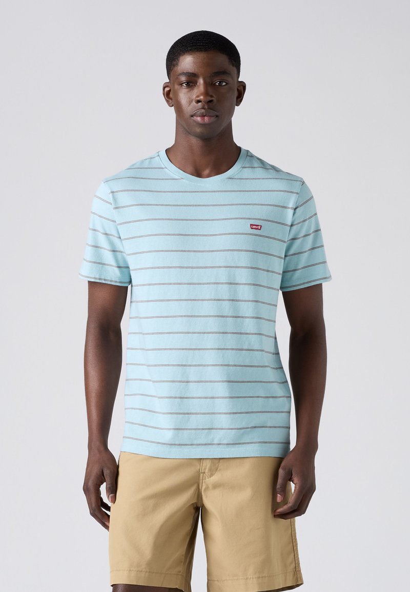 Hombre con camiseta de rayas azul claro y pantalones cortos beige, de pie frente a un fondo liso gris claro, mirando hacia adelante.