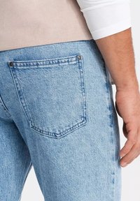 Vaqueros de denim azul claro con un diseño ajustado, que presentan un bolsillo trasero con remaches metálicos y costuras en contraste. Tejido texturizado con un ligero desgaste.