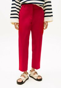 Pantalons droits rouges en tissu léger, présentant une texture lisse, avec une taille haute et des plis à l'avant. Associés à des sandales beiges.
