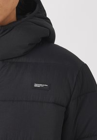 Svart puffjacka med en vadderad design, hög krage, justerbar huva och en varumärkesetikett på bröstet. Slätt, lättviktsmaterial.