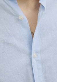 Camisa de lino azul claro con collar de botón, dos botones blancos y una tela texturizada con un ligero brillo.