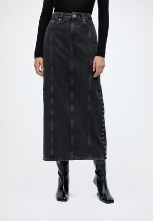 Person trägt einen hoch taillierten schwarzen Denim-Midirock mit bestickten seitlichen Details und schwarzen Stiefel mit Absatz vor weißem Hintergrund.