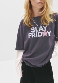 Grå oversized t-shirt med "SLAY FRIDAY" i hvid og et pink stjernedesign. Laget over en hvid langærmet skjorte. Blødt bomuldsstof.