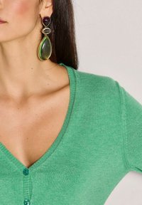 Groene cardigan met een V-hals en een knoopsluiting. Grote, multicolore oorringen met verschillende steenvormen en texturen.