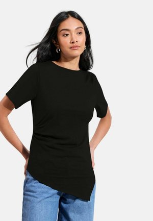 Kvinde med langt sort hår iført creoløreringe, en sort asymmetrisk kortærmet top og blå jeans, positurerende mod en hvid baggrund.