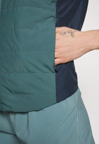Groene en marineblauwe pufferjack met een gequiltde textuur en een ritszak; gedragen met bijpassende lichtblauwe broeken.