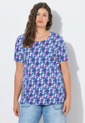 Frau mit langen, welligen Haaren, die ein blaues T-Shirt mit einem rosa-weißen Vogelmuster und hellblaue Jeans trägt, steht vor einem weißen Hintergrund.