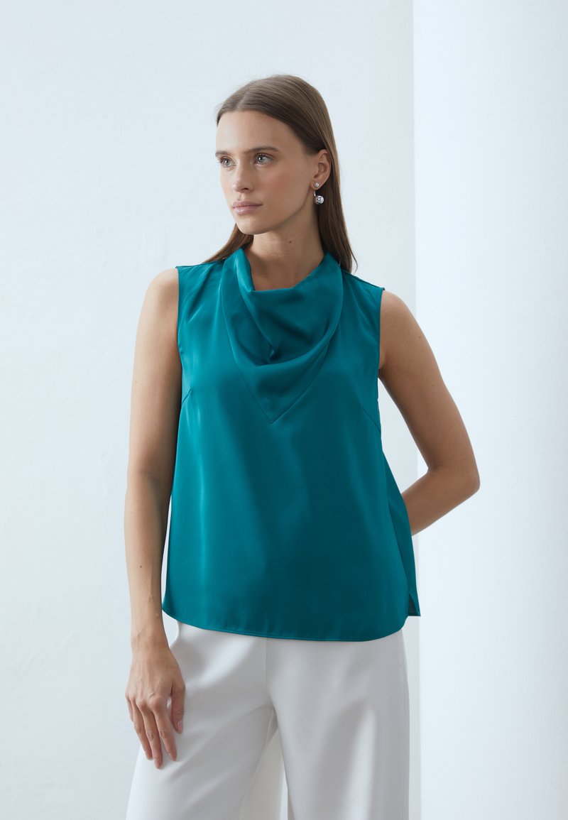 Top senza maniche in seta color teal con scollo drappeggiato, texture liscia, vestibilità morbida e fessure laterali, abbinato a pantaloni wide-leg bianchi.