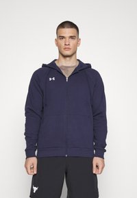 Marine blauwe zip-up hoodie met een voorklepzak, capuchon met trekkoord en geborduurd logo op de borst; gecombineerd met zwarte shorts met een logo.