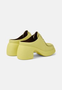Scarpe in pelle gialla con punta arrotondata, ampia suola e un design del tacco unico e curvato. Dettagli di cucitura lungo il bordo superiore.