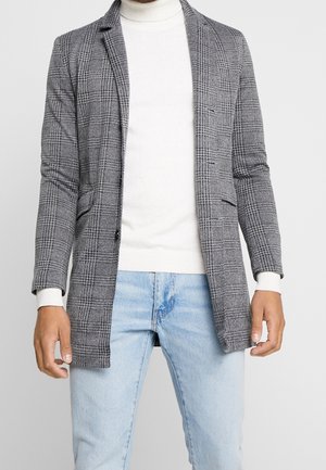 Mann trägt grauen karierten Blazer über einem weißen Rollkragenpullover und hellblauen Jeans, steht vor einem schlichten Hintergrund.