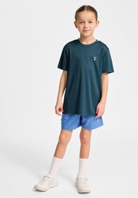 Mörkblågrön kortärmad t-shirt med en liten logotyp, kombinerad med ljusblå shorts. Vita strumpor och ljusa sneakers kompletterar outfiten.
