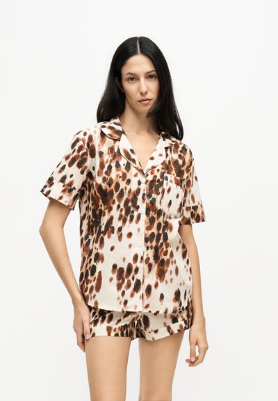 Femme aux longs cheveux foncés portant une chemise à manches courtes boutonnée et un short assorti avec un motif tacheté marron et noir sur fond blanc.
