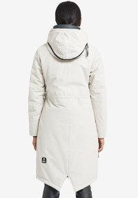 Valkoinen pitkä parka-takki hupulla, jossa on kiristettävä vyötärö, sileä rakenne ja brändilogo takana alhaalla.