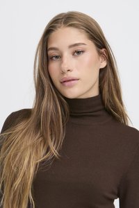 Jeune femme aux longs cheveux châtain clair portant un pull à col roulé marron foncé, regardant légèrement hors champ sur un fond uni.