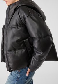 Black padded jacket with a hood, featuring a glossy faux leather material, snap button closures, and side pockets. Styled with blue jeans.