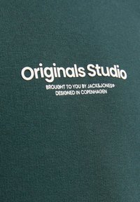 Tessuto di cotone verde con una texture liscia. Presenta un testo bianco: "Originals Studio" e "presentato da JACK & JONES, progettato a Copenaghen."
