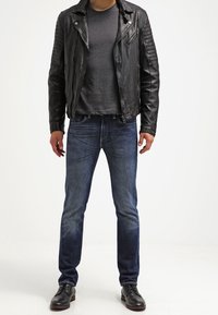 Sort lædermotorcykeljakke med strukturerede skuldre, grå t-shirt under, blå jeans og sorte lædersko. Enkel, tætsiddende design.