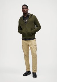 Cápsula verde-azeitona com fecho éclair por cima de uma camisa xadrez preto e branco, combinada com calças cargo em tom de camelo e sapatos pretos. Texturas suaves e design informal.