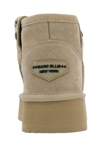 Stivale alla caviglia in suede beige con suola in gomma piatta, dotato di una toppa con logo e dettagli metallici ai lati. Finitura opaca texturizzata.