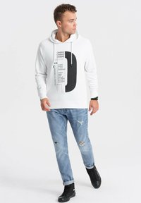 Sudadera blanca con diseño gráfico negro, cordones y puños acanalados, combinada con jeans rasgados de color azul claro y zapatillas altas negras.