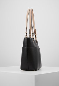 Musta nahkalaukku, jossa on säilytetty texture, kahdella pitkällä beige-nahkahihnalla ja kultaisilla yksityiskohdilla. Se seisoo valkoisella pinnalla.