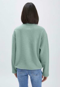 Pull oversized vert menthe en tissu texturé, avec des épaules tombantes et un détail de tag au dos. Porté avec un jean en denim bleu.