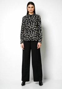 Schwarze Bluse mit einem weißen abstrakten Muster, mit Kragen, langen Ärmeln, kombiniert mit hoch sitzenden, schwarzen, weiten Hosen und Absatz-Stiefeletten.