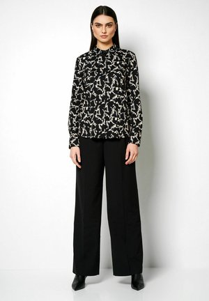 Schwarze Bluse mit einem weißen abstrakten Muster, mit Kragen, langen Ärmeln, kombiniert mit hoch sitzenden, schwarzen, weiten Hosen und Absatz-Stiefeletten.