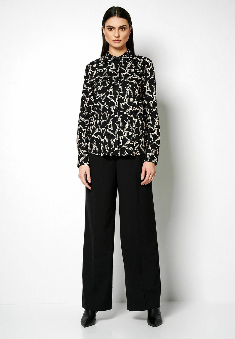 Schwarze Bluse mit einem weißen abstrakten Muster, mit Kragen, langen Ärmeln, kombiniert mit hoch sitzenden, schwarzen, weiten Hosen und Absatz-Stiefeletten.