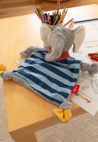 Peluche di elefante grigio con grandi orecchie, corpo di coperta a strisce blu, sciarpa arancione e piedi gialli, poggiato su un tavolo di legno accanto a matite colorate.