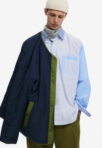 Padded jacket in navy with olive trim, paired with a blue and white striped shirt featuring a chest pocket. Model wears a beige beanie.