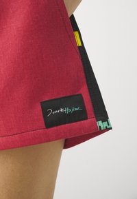 Røde stoffshorts med en kontrasterende svart sidepanel, med et logo-merke i svart med teal tekst og et gult aksent nær sømmen.