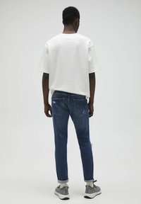 Man med vit oversized T-shirt, mörkblå upprullade jeans med slitna detaljer, vita strumpor och grå sneakers står mot en enkel bakgrund.