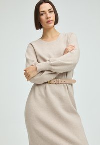 Beiges Strickkleid mit langen Ärmeln, Rundhalsausschnitt und lockerer Passform, tailliert mit einem hellrosa Gürtel mit goldener Schnalle.