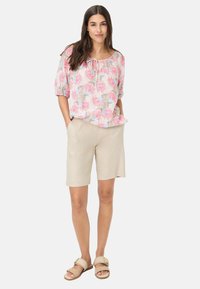 Blouse à imprimé floral avec des tons roses, bleus et beiges, manches courtes ; associée à un short beige et des sandales légères. Coupe décontractée, design casual.