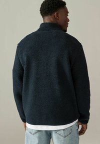 Pullover in fleece navy con colletto alto, superficie testurizzata e vestibilità rilassata. Camicia bianca a strati e jeans azzurri chiari visibili sotto.