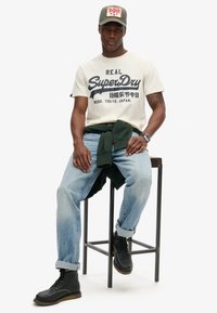Camiseta crema con texto de logo negro, jeans azul claro, sudadera verde atada a la cintura y botas negras con cordones, sentado en un taburete.