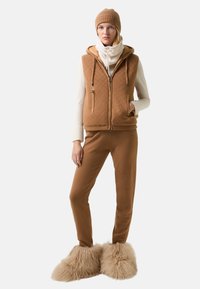 Brun polstret vest med hætte og to frontlommer, parret med matchende brune bukser og fuzzy beige fodtøj. Lagdelt creme strikket tørklæde.