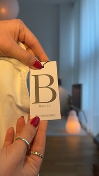 White ribbed fabric with a tag labeled "B Basics" by Stradivarius, attached by a black string. Hand holding the fabric with rings visible.