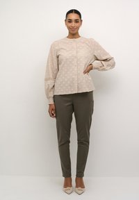 Chemise beige clair avec un motif à pois texturé, encolure ronde et fermeture à boutons. Associée à un pantalon olive ajusté et des chaussures crème.