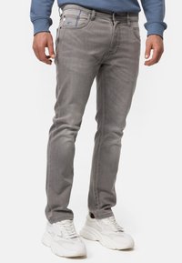 Jeans de mezclilla gris con un corte slim, que presentan un acento azul en el bolsillo delantero, combinados con zapatillas blancas. Textura suave y detalles de costura.