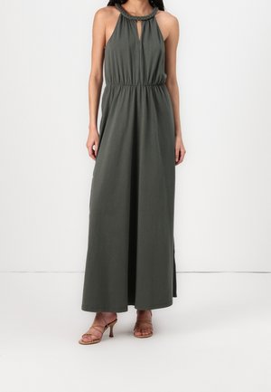 Mujer con vestido maxi verde oscuro sin mangas con escote halter trenzado y abertura frontal, combinado con sandalias de tacón marrón con tiras.