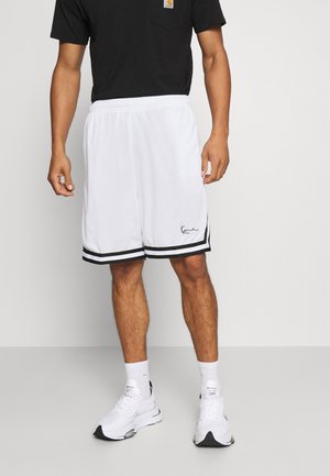 Hombre con pantalones cortos deportivos blancos con ribete negro y zapatillas blancas, de pie frente a un fondo claro y liso.