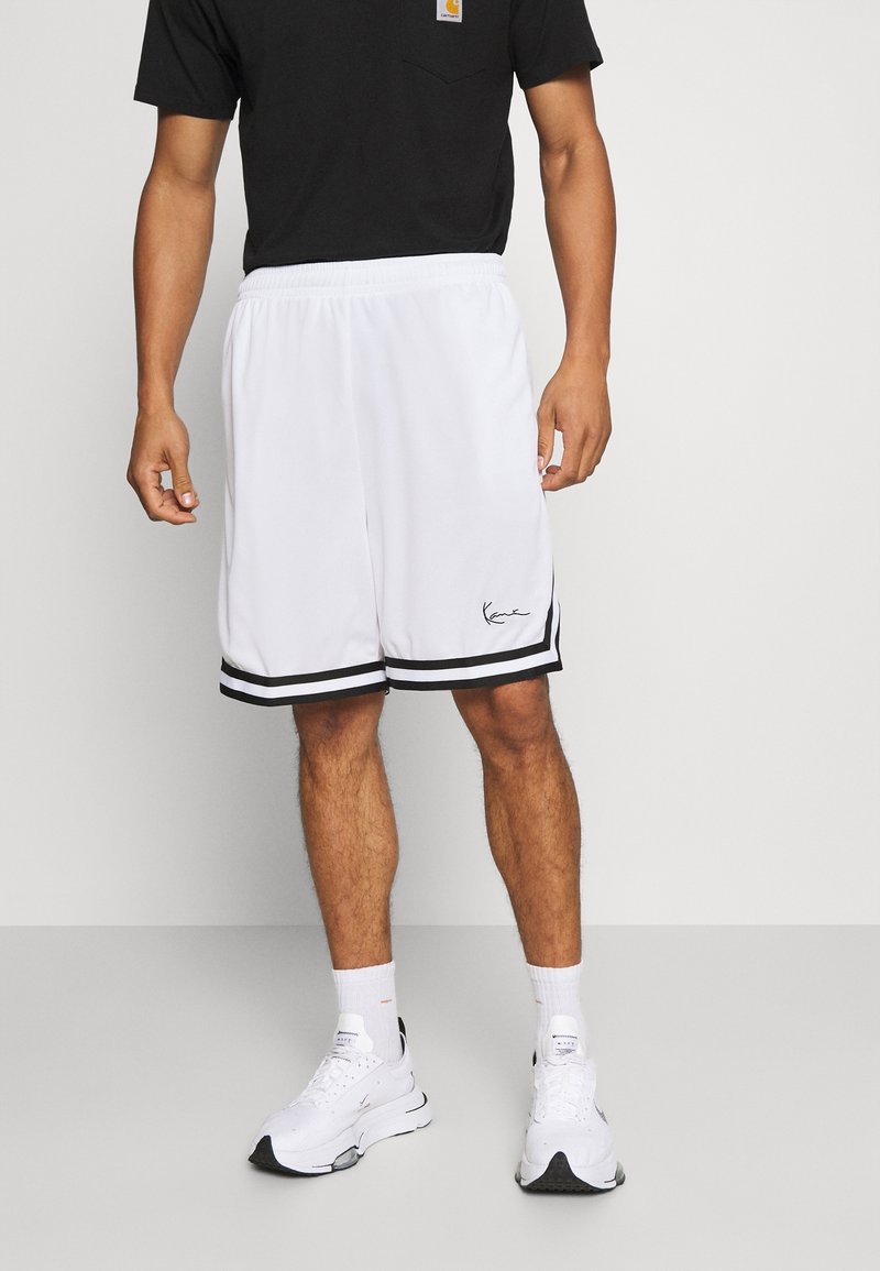 Homme portant un short de sport blanc avec une bordure noire et des baskets blanches, debout devant un fond clair uni.