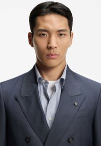 Giovane uomo con capelli corti neri che indossa una giacca doppiopetto blu scuro sopra una camicia a righe azzurra e una maglietta bianca sotto.