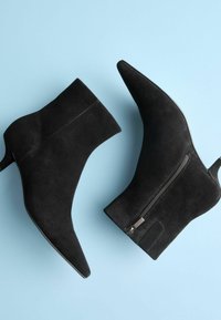 Čierne semišové členkové topánky s špicatou špičkou, bočným zipsom a elegantným opätkom, umiestnené na svetlom modrom pozadí.